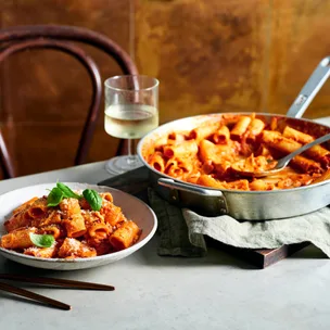 Plate of rigatoni pasta with tomato sauce and basil, a pan of pasta, and a glass of white wine on a table.