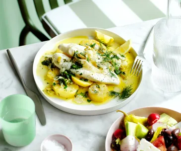 A fillet of white fish on a plate with white sauce and potatoes. There is a mint green background. 
