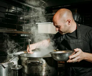 Mid-shot of a male chef in a black apron, stirring a large steel pot, with steam rising from it. 